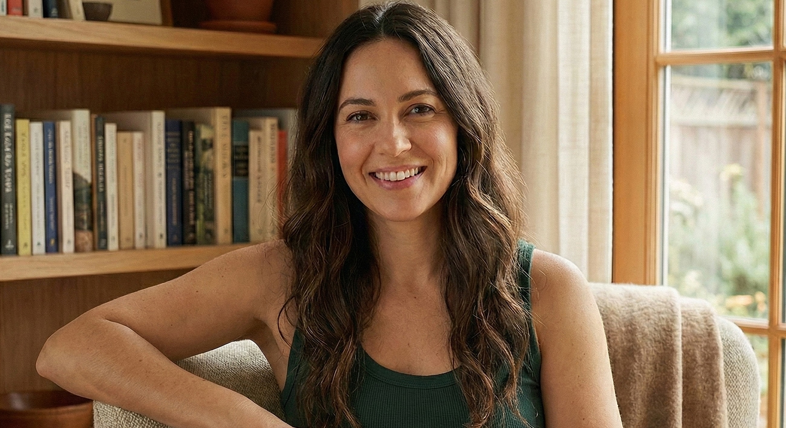 Smiling woman sitting by a window and bookshelf.